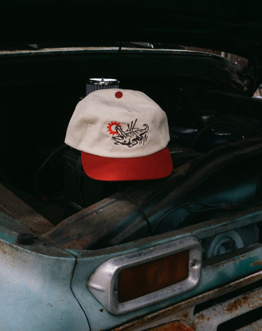 A cream and red baseball cap with a black and red graphic on the front, resting on the open hood of a rusted teal vintage truck.