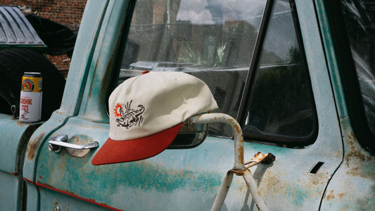 A cream and red baseball cap with a graphic print of an alligator and sun, hanging from a rusty metal mirror on the side of a weathered turquoise truck.