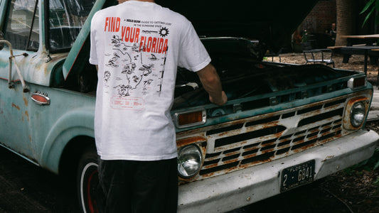 A person wearing a white graphic t-shirt with 'FIND YOUR FLORIDA' printed on the back, standing in front of a vintage, rusted teal pickup truck with the hood open.