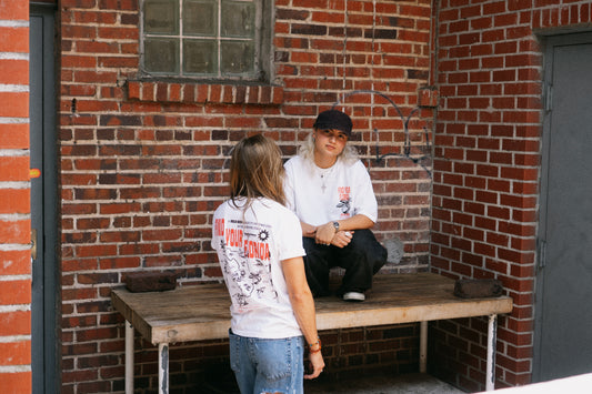 A person in a white graphic t-shirt and jeans stands in front of a brick wall, facing a person sitting on a wooden workbench wearing a white t-shirt, black pants, and a black cap.