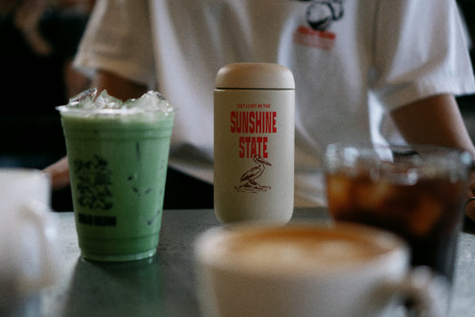 A beige insulated travel mug with red 'GET LOST IN THE SUNSHINE STATE' text and a bird illustration sits on a cafe table next to a green iced drink, with a person in a white shirt blurred in the background.