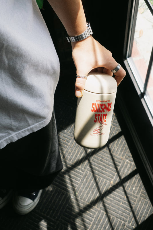 A person in a white t-shirt and black pants holds a cream-colored insulated tumbler with red 'GET LOST IN THE SUNSHINE STATE' text and a red pelican graphic, standing near a window with strong sunlight and shadows.