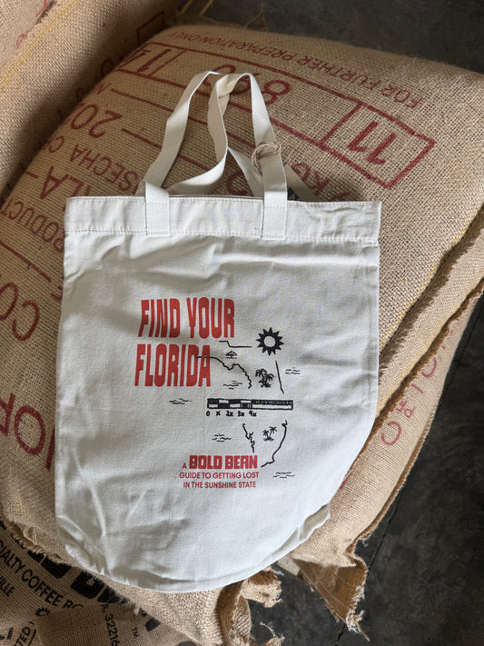 A white canvas tote bag with red and black graphic text reading 'FIND YOUR FLORIDA' and 'A BOLD BERN' lies on top of large brown burlap sacks.